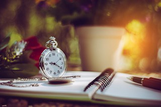 Image of desktop with pen, pocket watch and rose on an a spiral-bound notebook open to a blank sheet