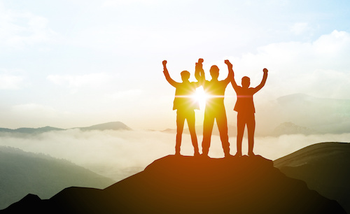 Image of team joining hands in triumph on a mountain peak