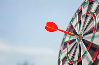 Image of a dart board with red-tipped dart in the bullseye with sky background