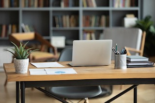 Image of a modern cozy workspace in a home: a maple work desk with laptop. documents, jar of pens, a wall of books, and comfortable seating