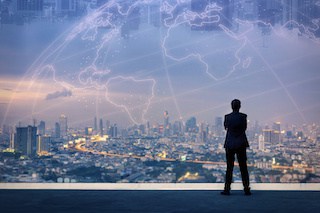 Image of businessman with his back to us looking thru a large glass window from a high floor at a cityscape over which the world map appears in faint cloud-like outline superimposed over a twilight sky