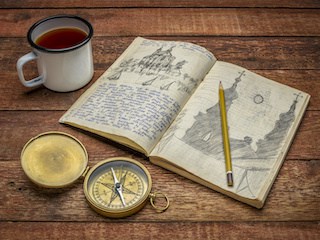 Image of a vintage travel journal with handwriting and pencil sketches, an old brass compass and a mug of tea on a rustic table