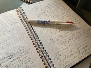 Image of a page of Jean's thinking notebook of handwritten thoughts with colored notes and a Thinking Directions pen laying atop the page