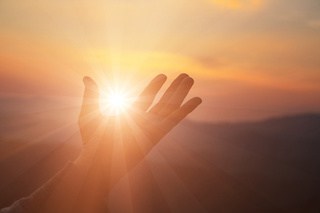 Image of a woman's hand raised as if holding the sun in her palm; sunset background.