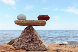 Image of white stone & red stone balancing each other atop a flat rock atop a pyramid-shaped rock, with the sea & sky in background