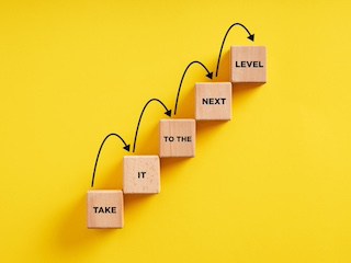 Image of small wooden block tiles arranged in a row rising upward like a staircase with the words Take-It-to the - Next-Level on them, centered against a golden yellow background with arrows stepping up from block to block.