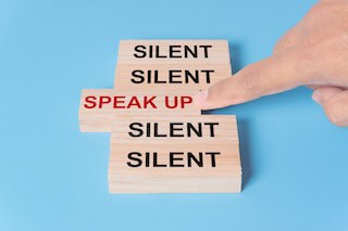 Image of Finger pushing forward a wooden block that says "Speak Up," which is sandwiched between four blocks that each say "Silent."