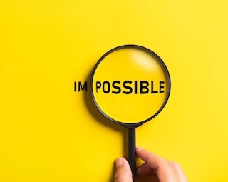 Image of a right hand holding a magnifying glass enlarging "Possible" of the word Impossible, printed in black on sunny yellow background