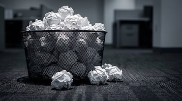 Image of an overflowing wire mesh trash bin filled with crumpled paper balls on an office floor